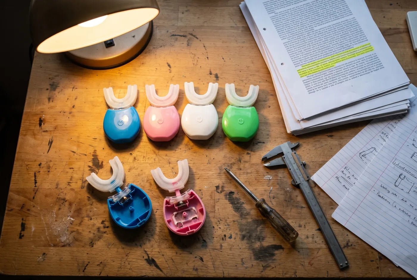 A workbench with several taken-apart U-shaped toothbrushes beside printed research papers